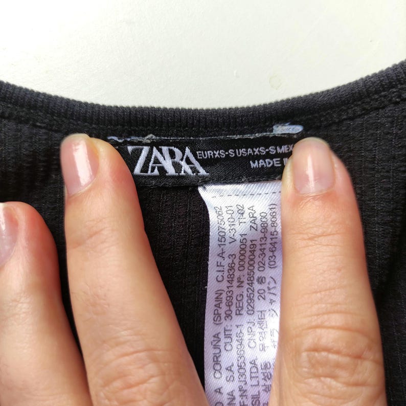 Puede incluir: Primer plano de una prenda negra de tejido acanalado con una etiqueta ZARA. La etiqueta muestra texto como "EURXS-S USAXS-S MEX" y otros detalles. La prenda est&aacute; siendo sostenida por una persona.