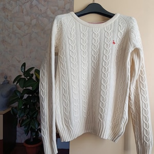 May include: Cream-colored cable knit sweater with long sleeves. The sweater features a small red emblem on the chest and a ribbed hem and cuffs. The sweater is hanging on a black hanger.