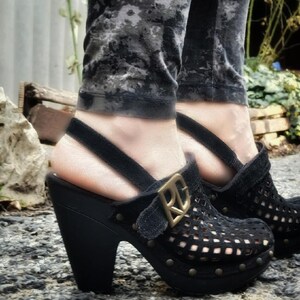 May include: Black suede clogs with a gold buckle and a woven design. The clogs have a high, chunky heel.