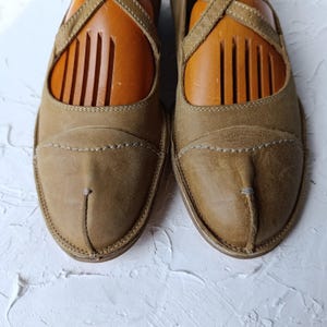 May include: A pair of tan leather shoes with a unique design. The shoes have a rounded toe and a strap across the top. The shoes are filled with wooden shoe trees. The shoes are on a white textured surface.
