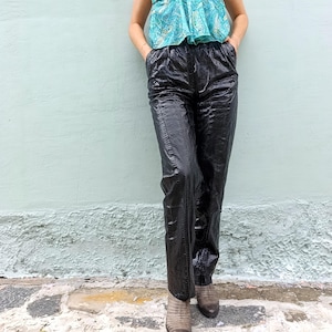 May include: High-waisted black trousers featuring a glossy, textured snakeskin-like pattern. These pants have a straight-leg silhouette, an elasticized waistband, and side pockets. The material has a reflective sheen, highlighting the intricate scale-like surface texture throughout the garment.