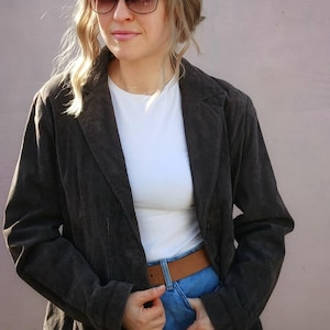May include: A brown suede blazer with a white t-shirt underneath and blue jeans. The blazer has a notched collar and long sleeves. The person is wearing a brown belt.