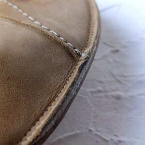 May include: Close-up of a tan leather shoe's edge. The shoe features white stitching along the seams and a textured surface. The sole shows signs of wear and tear, with visible separation.