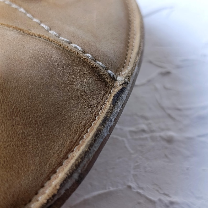May include: Close-up of a tan leather shoe's edge. The shoe features white stitching along the seams and a textured surface. The sole shows signs of wear and tear, with visible separation.