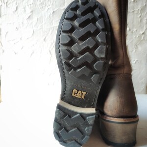 Może przedstawiać: Brązowe sk&oacute;rzane buty z czarną gumową podeszwą. Podeszwa ma bieżnik i napis "Caterpillar" nadrukowany na niej.