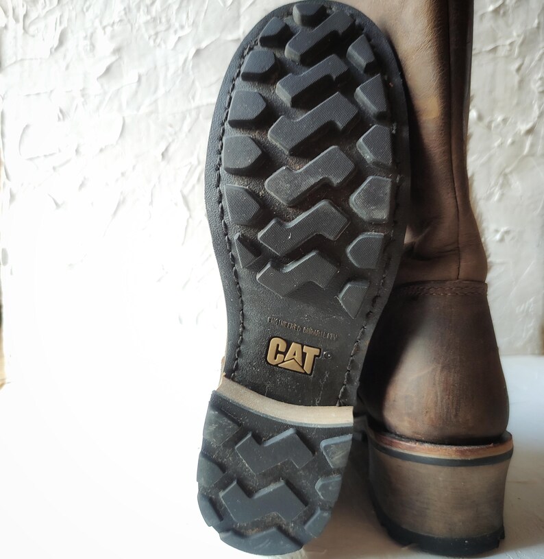 Może przedstawiać: Brązowe sk&oacute;rzane buty z czarną gumową podeszwą. Podeszwa ma bieżnik i napis "Caterpillar" nadrukowany na niej.