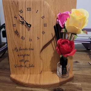 May include: A light brown wooden clock with a curved edge and black Roman numerals. The clock face features the engraved text: My favourite time is anytime with you. Loretta & Julia. A small glass diffuser with pink, yellow, and red artificial roses.
