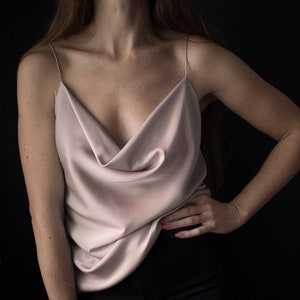 May include: A pale pink silk camisole with a cowl neck and thin straps. The top is worn with black trousers. The image is set against a dark backdrop, emphasising the smooth texture and flow of the material.