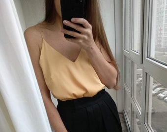 May include: A woman is wearing a light peach colored satin tank top and black high-waisted pants. The pants have pleats in the front.