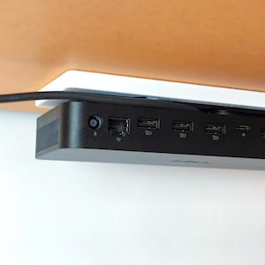 Könnte beinhalten: Eine schwarze Dell-Dockingstation mit mehreren Anschlüssen, montiert unter einer braunen Oberfläche. Die Dockingstation verfügt über verschiedene USB-Anschlüsse, einen Ethernet-Anschluss und einen Stromeingang. Eine weiße Montageplatte ist sichtbar.