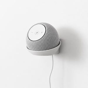 Può includere: Uno smart speaker sferico bianco con un esterno in tessuto a rete e un pannello superiore circolare piatto con un segno meno, un segno più e un'icona centrale. Il dispositivo è montato in un supporto bianco fissato a una parete con un cavo.