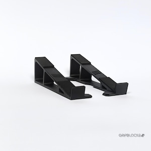 Op de afbeelding: Twee zwarte, schuine steunblokken met een getextureerd, antislip oppervlak. De blokken zijn ontworpen om een object te verhogen. De merknaam, GRIPBLOCKS, is rechtsonder in grijs gedrukt.