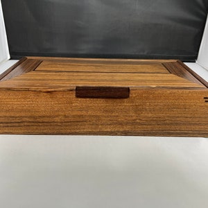 May include: A wooden box with a dark brown wood grain finish. The box has a simple, rectangular shape with a small, rectangular handle on the top.