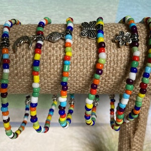 May include: Colorful beaded bracelets with silver charms, including a dolphin, fish, butterfly, flower, and heart. The bracelets are displayed on a burlap-covered stand, showcasing a variety of bright colors and unique designs.