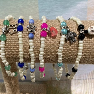 May include: Set of six white beaded bracelets with various charms, including a paw print, a flower, a heart, a turtle, and a skull. The bracelets are displayed on a brown burlap background.