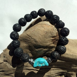 Pode incluir: Um bracelete de contas de rocha vulcânica preta com um amuleto de elefante turquesa. O bracelete está em um pedaço de madeira à deriva.