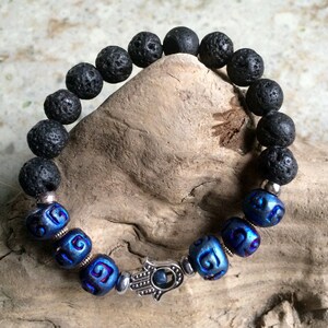 May include: A black lava rock bracelet with blue and silver beads. The bracelet features a silver hamsa hand charm.