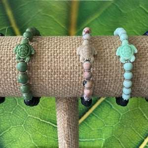 Puede incluir: Cinco pulseras con dijes de tortuga de cerámica. Las pulseras están hechas con cuentas de diferentes colores, incluyendo azul, verde, rosa y blanco. Las pulseras se muestran en un soporte de arpillera marrón.