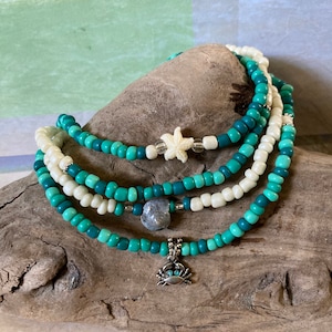 May include: Four layered beaded bracelets with a white, teal, and turquoise colour gradient. The bracelets feature a starfish charm, a grey stone charm, and a silver crab charm.