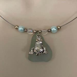 Peut inclure: Un pendentif en verre de mer bleu clair avec un charme de grenouille argentée suspendu à un collier en chaîne argentée. Le collier est accentué de perles bleu clair et transparentes.