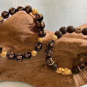 May include: Two bracelets with black beads and gold accents. One bracelet has black onyx beads with gold lettering and the other has black lava rock beads with a gold charm.