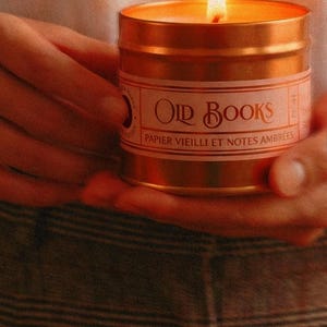 May include: A gold-coloured metal candle tin with a lit wick and a white label featuring the text "OLD BOOKS" and "PAPIER VIEILLI ET NOTES AMBR&Eacute;ES" in a decorative font. The candle emits a warm glow from the flame, illuminating the metallic surface.