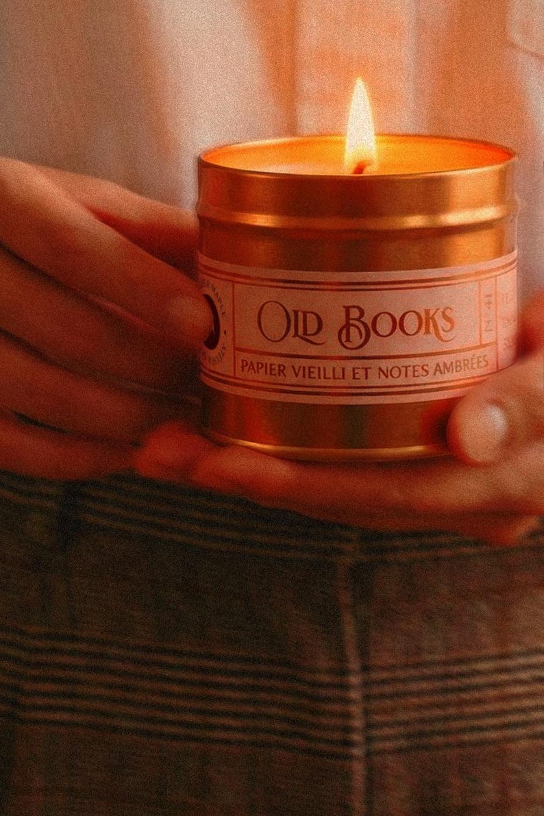 May include: A gold-coloured metal candle tin with a lit wick and a white label featuring the text "OLD BOOKS" and "PAPIER VIEILLI ET NOTES AMBR&Eacute;ES" in a decorative font. The candle emits a warm glow from the flame, illuminating the metallic surface.