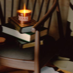 May include: A lit candle in a copper-coloured metal tin with a label reading "Old Books" resting on a stack of three hardcover books. The books are positioned on a dark wooden chair with a curved backrest and thin vertical spindles.