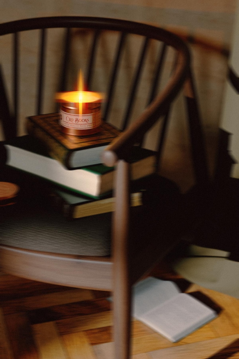 May include: A lit candle in a copper-coloured metal tin with a label reading "Old Books" resting on a stack of three hardcover books. The books are positioned on a dark wooden chair with a curved backrest and thin vertical spindles.