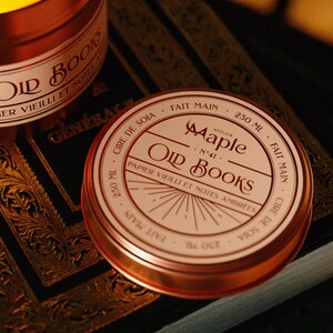 May include: A round copper-coloured metal candle tin with a white label featuring the text Atelier Maple No 41 Old Books, Papier Vieilli et Notes Ambr&eacute;es, 250 ml, Cire de Soja, and Fait Main. The lid displays a sunburst graphic design.
