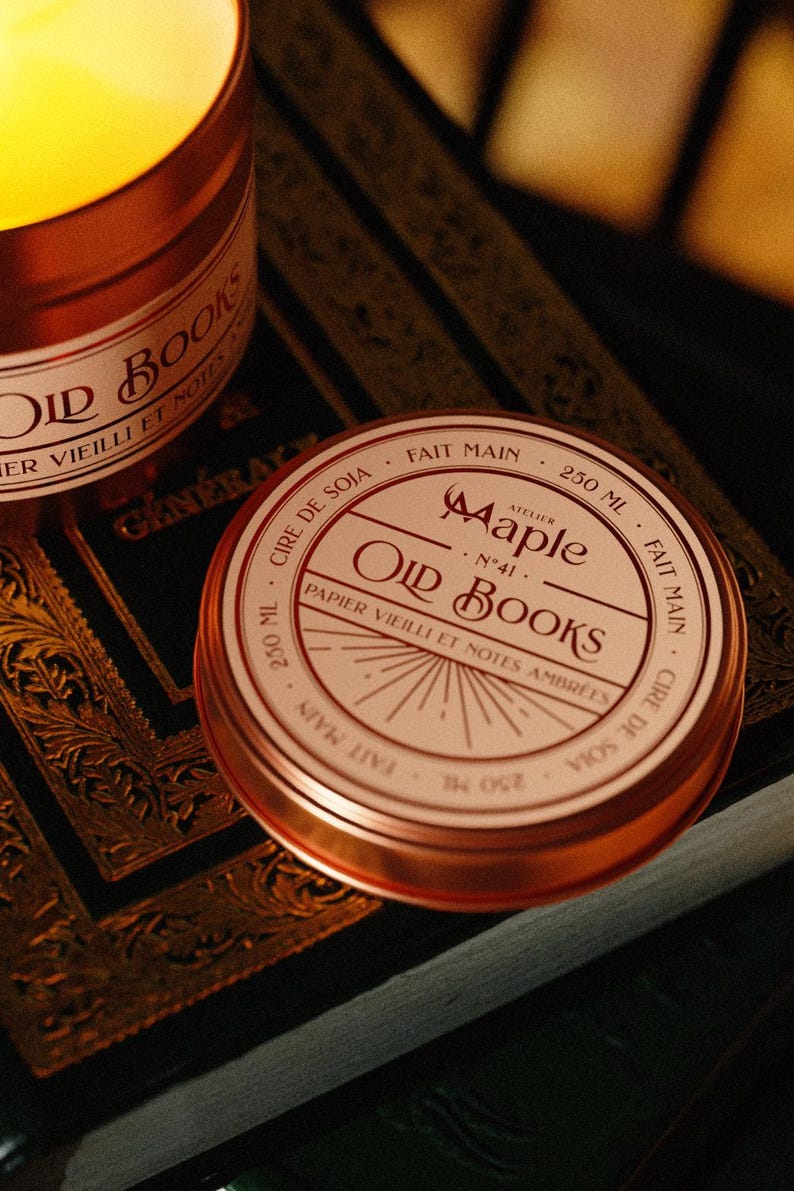 May include: A round copper-coloured metal candle tin with a white label featuring the text Atelier Maple No 41 Old Books, Papier Vieilli et Notes Ambr&eacute;es, 250 ml, Cire de Soja, and Fait Main. The lid displays a sunburst graphic design.