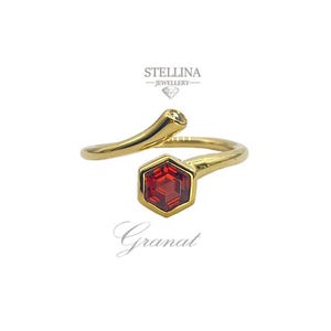 Può includere: Un anello dorato con una gemma rossa esagonale e una piccola pietra rotonda trasparente. L'anello ha un design curvo unico. La parola "Granat" è scritta in corsivo sotto.