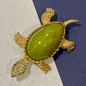 May include: A gold turtle brooch with a green stone cabochon in the center. The turtle is decorated with rhinestones.