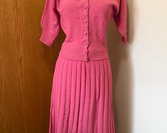 May include: A vintage pink knit two-piece set. The top is a button-down cardigan with short sleeves and embroidered detailing on the shoulders. The skirt is a pleated midi-length skirt. The set is displayed on a mannequin.