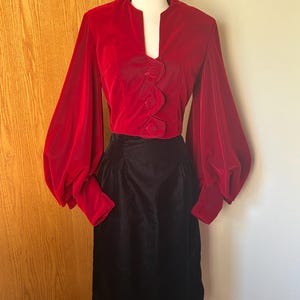 Può includere: Un top vintage in velluto rosso con maniche ampie e una gonna nera. Il top ha uno scollo a V e tre bottoni decorativi. La gonna è lunga fino al ginocchio ed è realizzata in un tessuto nero lucido. L'outfit è esposto su un manichino.
