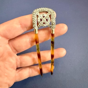 Peut inclure: Un peigne à cheveux Art Déco avec un dessus rectangulaire argenté incrusté de petits strass transparents. Le centre présente un motif décoratif. Les deux dents ont un motif écaille de tortue brun, ambre et vert.