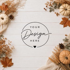 May include: A white wooden surface featuring a central black circular frame with the text YOUR Design HERE and a small heart icon. The scene is decorated with cream-colored fabric, orange autumn leaves, dried wheat, pinecones, and pumpkins.