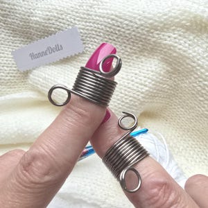 Könnte beinhalten: Zwei silberfarbene Garnspannringe aus Metall, jeweils mit einem gewickelten Drahtdesign und zwei kleinen Schlaufen an den Enden. Die Ringe werden an den Fingern getragen, im Hintergrund ist ein weißes Etikett mit dem Text HanneDolls zu sehen.