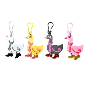Op de afbeelding: Vier eendvormige sleutelhangers met plastic clips. De eenden zijn wit, geel, zwart en roze, elk met een sjaal met stippen en glinsterende laarzen. De witte eend heeft zwarte laarzen, de gele roze, de zwarte rode en de roze eend heeft gouden laarzen.