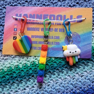 Pode incluir: Três marcadores de ponto de cores arco-íris com um coração, uma pilha de blocos de Lego e uma nuvem com um arco-íris. Os marcadores de ponto estão presos a um cordão branco com um cartão branco atrás. O cartão tem o texto "HANNFOOLS HANDMADE ITEMS FOR YOU".