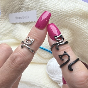 Könnte beinhalten: Drei verstellbare Metallringe mit einem stilisierten Katzensilhouetten-Design. Ein Ring ist silberfarben, während zwei Ringe schwarz sind. Die Ringe sind offen gestaltet, um eine Größenanpassung zu ermöglichen. Ein weißes Etikett mit HanneDolls ist sichtbar.