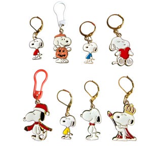 Op de afbeelding: Een verzameling van acht metalen bedels met het personage Snoopy in verschillende outfits en houdingen, waaronder een pompoenkostuum, een kerstmanpak en een kroon. De bedels zijn bevestigd aan goudkleurige oorbelhaakjes of plastic lussen.