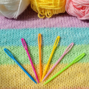 Può includere: Una collezione di filati colorati e aghi da maglia in plastica. Gli aghi sono blu, rosa, arancioni, gialli e verdi. Il filato è bianco, giallo e rosa. Lo sfondo è un tessuto a maglia arcobaleno.