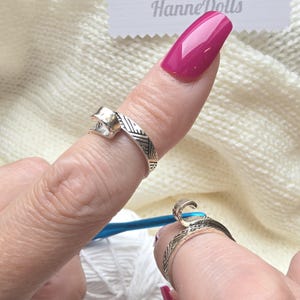 Op de afbeelding: Twee zilverkleurige metalen garenspanningsringen met een open, verstelbaar bandontwerp en een gestructureerd, geometrisch patroon op de buitenkant. Elke ring heeft een kleine, gebogen haak aan de bovenkant voor het geleiden van garen tijdens het haken.