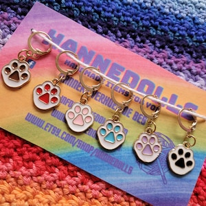 Op de afbeelding: Set van zeven gouden steekmarkeerders met pootjescharms. De charms zijn wit met verschillende gekleurde pootjesafdrukken: bruin, rood, roze, blauw, paars en zwart.