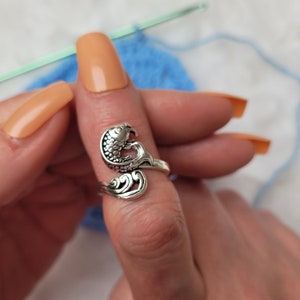 Op de afbeelding: Een zilveren ring met een gedetailleerd ontwerp van een vis die in golven zwemt. De ring wordt gedragen op een vinger met lichtbruine nagellak.
