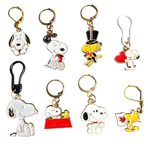 Op de afbeelding: Een verzameling van acht metalen bedels met Snoopy- en Woodstock-figuren in verschillende houdingen, waaronder een met een hoge hoed, een met een rood hart en een met een rood hondenhok. Elke bedel is bevestigd aan een goudkleurige of kunststof sluiting.