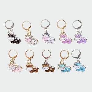 Pode incluir: Um conjunto de dez brincos de argola de metal, cada um com um pequeno pingente em forma de vaca com manchas em forma de coração. Os pingentes vêm em várias cores, incluindo preto, branco, rosa, roxo, marrom e azul, com argolas em tons de prata ou ouro.