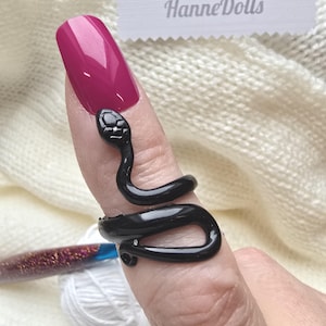 Può includere: Un anello nero a forma di serpente avvolto attorno a un dito con un'unghia color magenta. L'anello presenta una finitura liscia e lucida e un design dettagliato della testa. Il testo HanneDolls è visibile nella parte superiore dell'immagine.