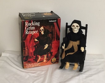 May include: A Halloween decoration featuring a skeletal Grim Reaper in a black robe, seated in a black rocking chair. The figure has a white skull face and is next to a box with the words "Rocking Grim Reaper" and an illustration of the figure.